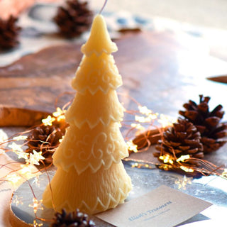 A small decorated beeswax candle shaped like a Christmas tree, placed on a table with pine cones and Christmas lights in the background.