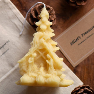 A beeswax candle shaped like a Christmas tree with a nativity scene, placed on a wooden surface with pine cones and a business card in the background.